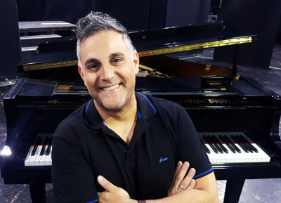 יוסף ארידן כשפניו למצלמה משלב ידיים ומחייך וברקע פסנתר כנף בסטודיו לפיתוח קול