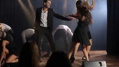 גבר ואשה בבגדי ערב רוקדים על במה מאחוריהם רקדנים ולפניהם קהל