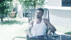 יוני סופר יושב על כסא בחצר בית ביום, מאחוריו שני עצים